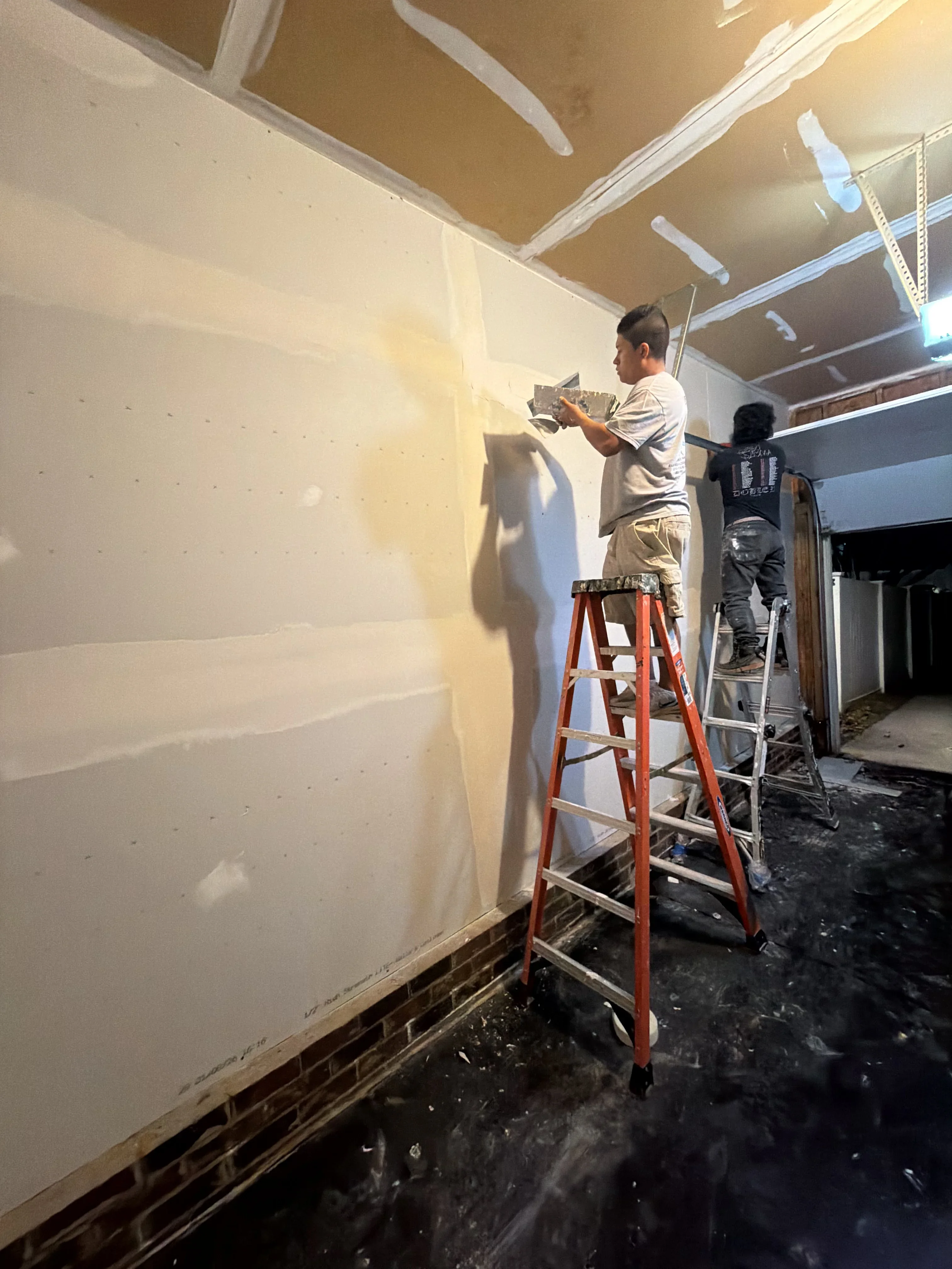 Worker on ladder applying drywall joint compound and tape to interior wall seams